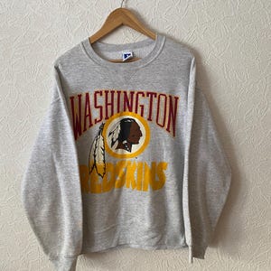 Pode incluir: Uma camisola de gola redonda cinzenta mesclada com o texto WASHINGTON em letras vermelho-escuras sobre um logótipo circular amarelo com uma ilustração de perfil de uma figura nativa americana e duas penas, com a palavra REDSKINS em amarelo abaixo.