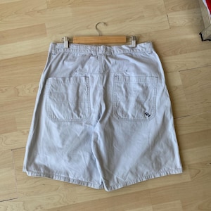 May include: Khaki-colored shorts with two back pockets and a small logo on the right pocket. The shorts are hanging on a wooden hanger. The shorts are made of a durable fabric and have a relaxed fit.