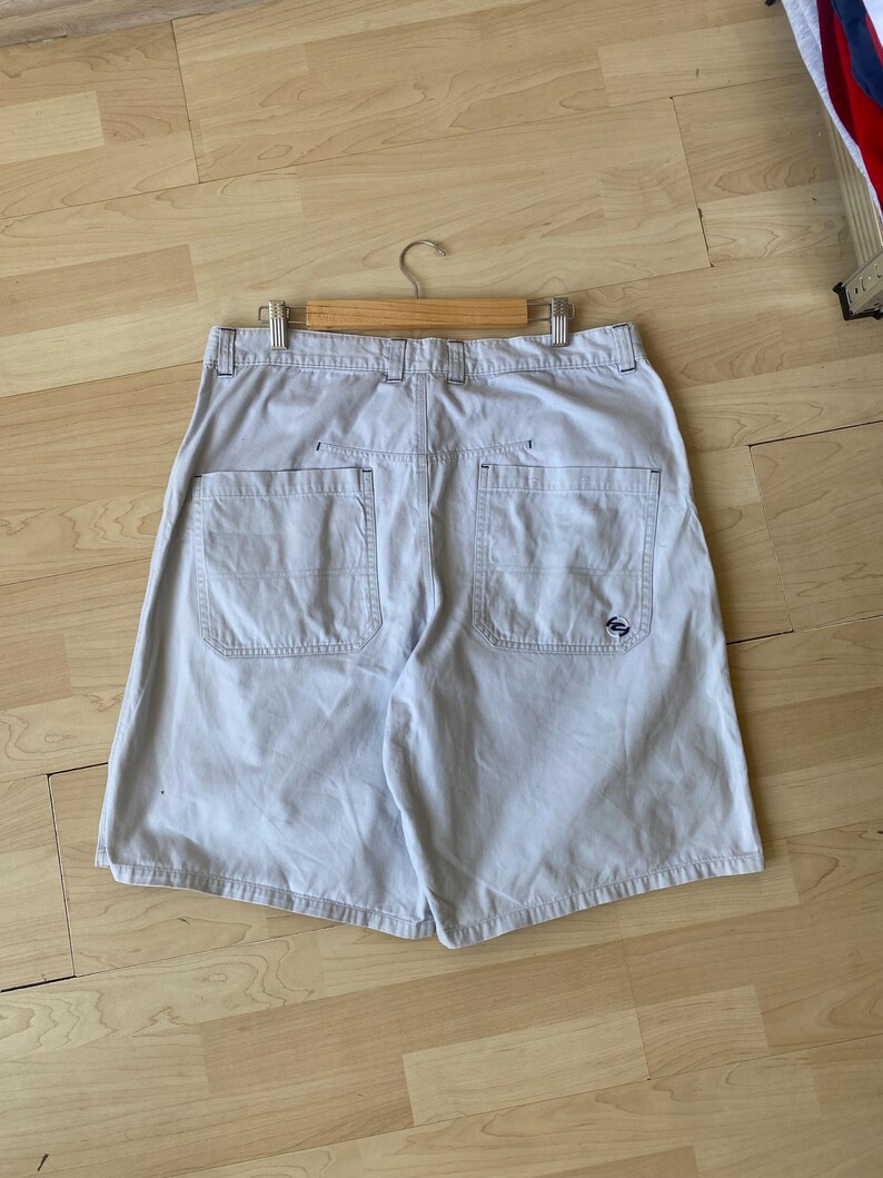 May include: Khaki-colored shorts with two back pockets and a small logo on the right pocket. The shorts are hanging on a wooden hanger. The shorts are made of a durable fabric and have a relaxed fit.