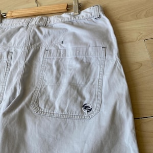 May include: Light beige cargo shorts with multiple pockets and a small navy blue logo on the back pocket. The shorts are hanging on a wooden hanger.