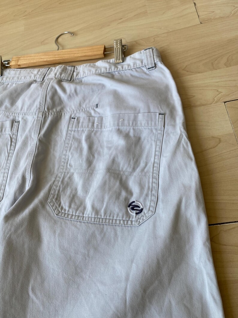May include: Light beige cargo shorts with multiple pockets and a small navy blue logo on the back pocket. The shorts are hanging on a wooden hanger.