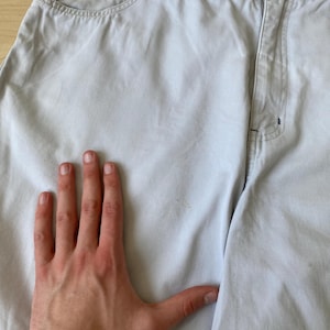 May include: Light gray denim shorts with a button closure and pockets. The shorts have a relaxed fit and a faded appearance. The brand name is visible on the waistband. A hand is placed on the shorts.