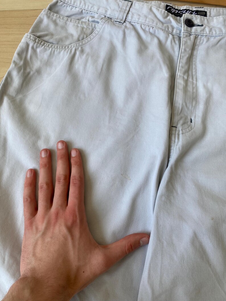 May include: Light gray denim shorts with a button closure and pockets. The shorts have a relaxed fit and a faded appearance. The brand name is visible on the waistband. A hand is placed on the shorts.