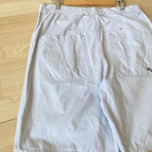 May include: Light blue shorts with two back pockets and a small logo on the right pocket. The shorts are hanging on a wooden hanger. The shorts are made of a light-weight fabric.