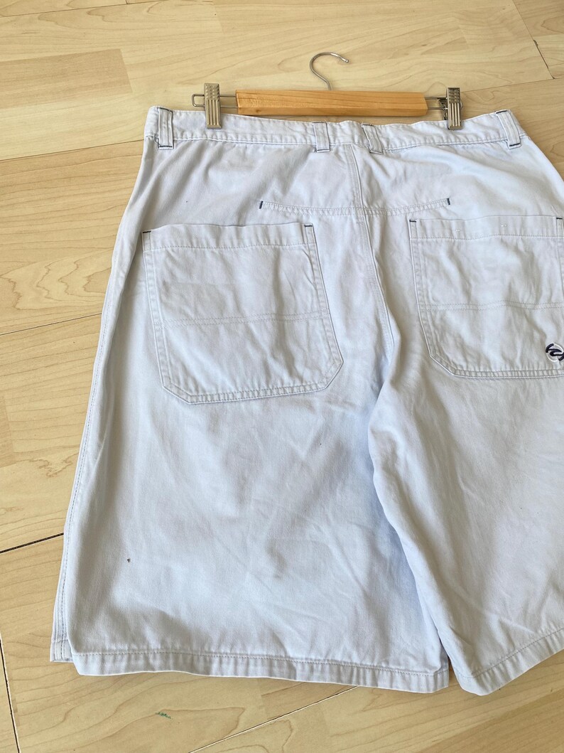 May include: Light blue shorts with two back pockets and a small logo on the right pocket. The shorts are hanging on a wooden hanger. The shorts are made of a light-weight fabric.
