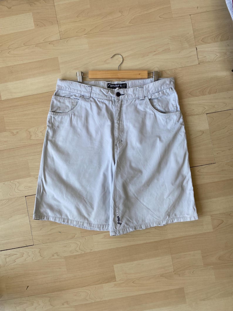 May include: Light gray shorts with a button closure and front and back pockets. The shorts are hanging on a wooden hanger. The brand name "Rip Curl" is visible on the waistband.