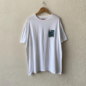 May include: White short-sleeved t-shirt with a small square graphic on the chest pocket. The graphic features a colourful abstract design with the word "ARENA" in white. The shirt is hanging on a wooden hanger.