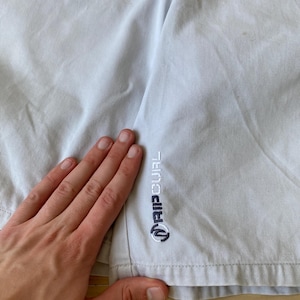 May include: Light gray shorts with the "RIP CURL" logo embroidered in navy blue. The shorts are made of a light fabric and appear to be slightly wrinkled. The logo is located on the lower left side of the shorts.