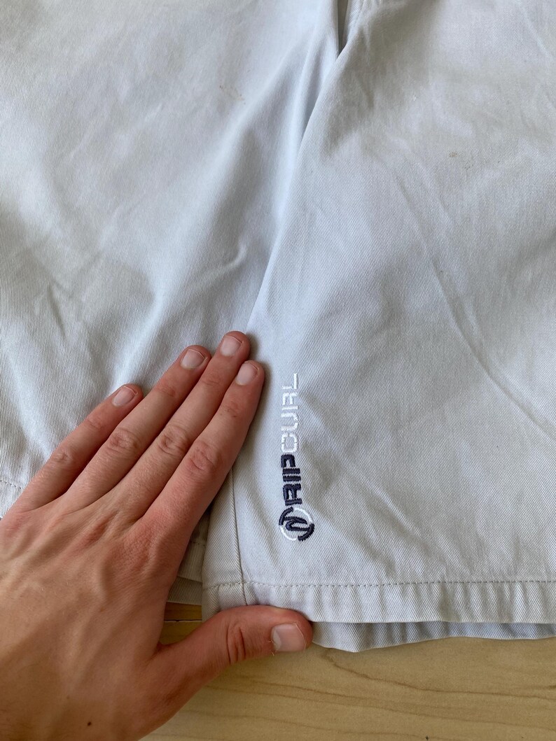 May include: Light gray shorts with the "RIP CURL" logo embroidered in navy blue. The shorts are made of a light fabric and appear to be slightly wrinkled. The logo is located on the lower left side of the shorts.
