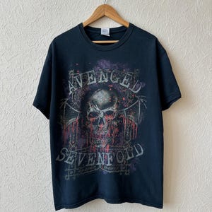Puede incluir: Una camiseta negra de manga corta con un estampado gráfico de una calavera con alas de murciélago y salpicaduras de pintura roja. El texto "AVENGED" está impreso en arco sobre la calavera y "SEVENFOLD" en línea recta debajo de ella.
