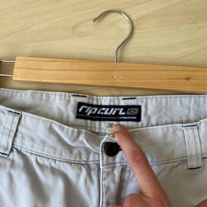 May include: Light gray shorts with a button closure and belt loops, hanging on a wooden hanger. The shorts have a size 36 label. The brand name "Rip Curl" is visible on a tag.