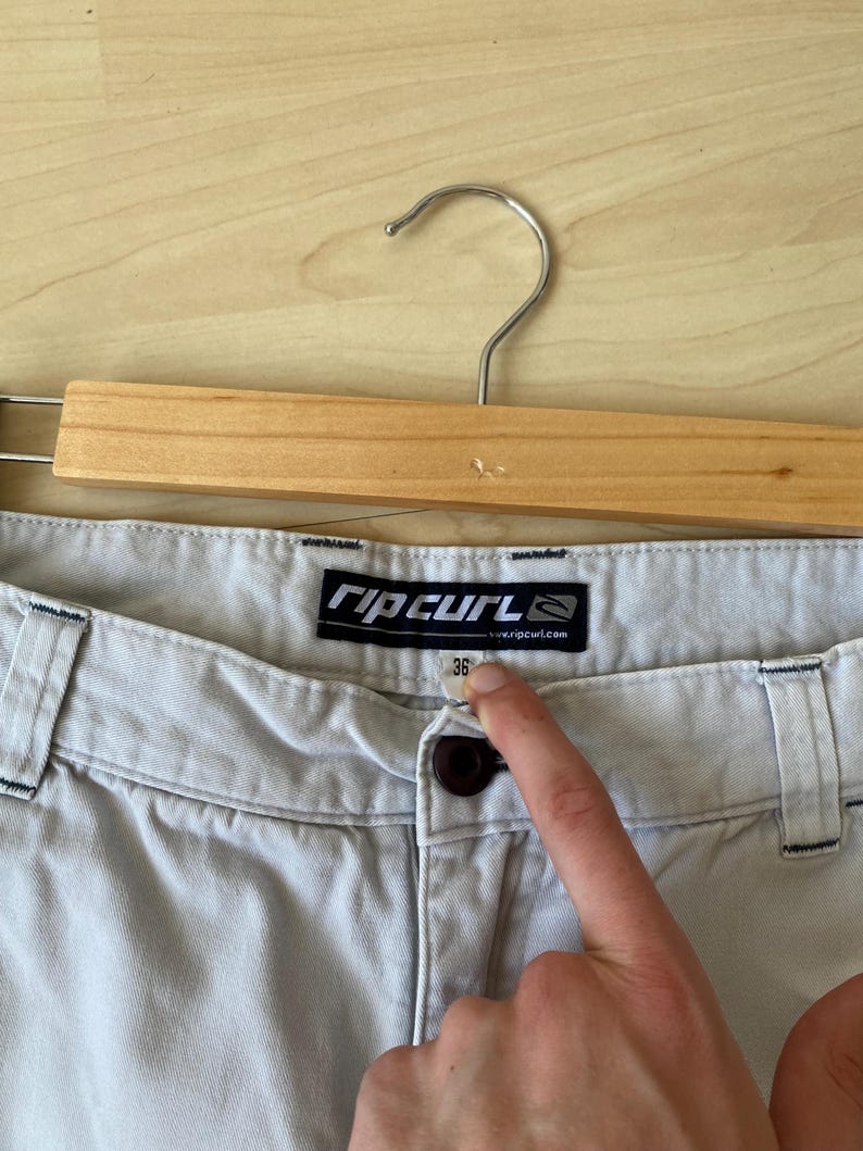 May include: Light gray shorts with a button closure and belt loops, hanging on a wooden hanger. The shorts have a size 36 label. The brand name "Rip Curl" is visible on a tag.