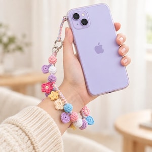 Pode incluir: Um smartphone violeta claro com capa transparente preso a uma alça de pulso de crochê feita à mão. A alça apresenta um cordão trançado adornado com várias formas de crochê, incluindo pequenas flores, estrelas e esferas em tons pastel.
