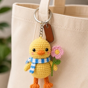 May include: A yellow crochet duck keychain featuring a blue and white striped scarf, orange feet, and a pink flower with a green stem. The duck has black eyes and pink cheeks, attached to a silver metal keyring with a brown leather-like tag.