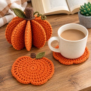 Puede incluir: Una calabaza naranja de ganchillo en tres dimensiones con un tallo marrón, una hoja verde y un zarcillo, acompañada de un posavasos plano, redondo y naranja de ganchillo con forma de calabaza bajo una taza de cerámica blanca con bebida.