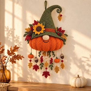 Peut inclure: Une décoration murale en crochet représentant un gnome en forme de citrouille avec un grand chapeau vert, orné d'un tournesol, de feuilles rouges et jaunes, et de pommes de pin. Des éléments suspendus incluent des mini-citrouilles.
