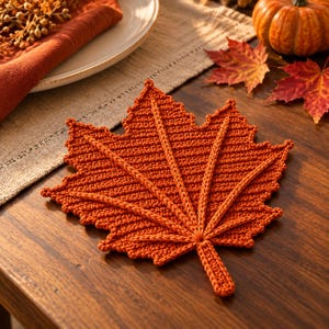 May include: A burnt orange crochet coaster shaped like a maple leaf. The textile piece features a detailed central vein structure radiating from the stem to the pointed lobes of the leaf, with a textured, ribbed stitch pattern throughout the surface.