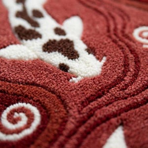 May include: A tufted textile piece featuring a white koi fish with brown spots swimming through a deep red background. The surface displays a textured, looped pile construction with swirling patterns that mimic water currents and ripples.