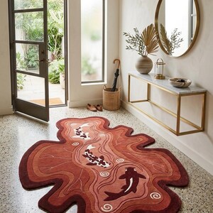 May include: An irregularly shaped area rug featuring a layered, wavy design in shades of terracotta, rust, and deep red. The rug depicts three koi fish swimming in a stylized pond pattern with white and dark brown accents and small spiral details.