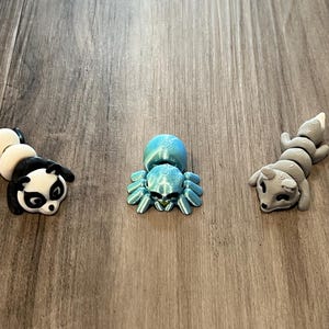 May include: Three small articulated figurines: a black and white panda, a metallic blue spider with a rounded abdomen, and a solid gray fox. Each figure features a segmented body design with visible 3D printed layer lines on the surface.