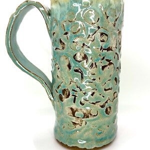 May include: A tall, handmade ceramic mug with a textured surface and a large handle. The mug features a turquoise glaze with brown and white accents. The rim has a brown glaze. The mug is suitable for hot or cold beverages.