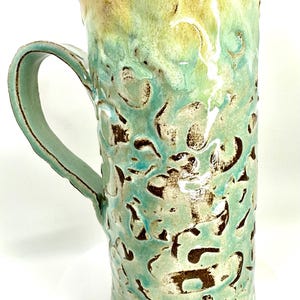 May include: Tall, handmade ceramic mug with a textured surface and a large handle. The mug features a blend of turquoise, green, and brown colors, with a glossy finish. The mug is approximately 6 inches tall.