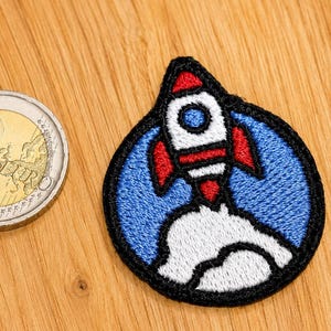 Puede incluir: Un parche bordado circular que muestra un cohete blanco con aletas rojas y una ventanilla azul, despegando de una nube blanca sobre un fondo azul, colocado junto a una moneda de 2 euros para mostrar su pequeño tamaño.