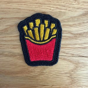 Könnte beinhalten: Ein gestickter Aufnäher, der einen roten Behälter gefüllt mit gelben Pommes frites zeigt. Das Design ist mit einem dicken schwarzen Rand umrandet, und das gesamte Stück ist mit einem strukturierten Faden vernäht, was eine klare Grafik ergibt.