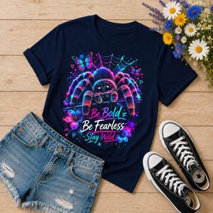 K&ouml;nnte beinhalten: Marineblaues T-Shirt mit einem farbenfrohen Spinnen-Design und dem Text "Be Bold, Be Fearless, Stay Wild". Das Design zeigt Schmetterlinge und Blumen. Schwarze und wei&szlig;e Turnschuhe und Jeansshorts sind ebenfalls im Bild.