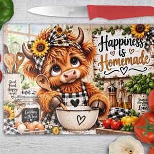 Peut inclure: Une planche à découper rectangulaire en verre illustrée d'une vache highland brune et poilue portant un nœud et un tablier à carreaux noirs et blancs, tenant une cuillère en bois au-dessus d'un bol. Le texte indique Happiness is Homemade, Good Food Happy People, Cooking with love et Season everything with Love.