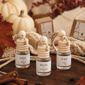 May include: Three small glass car air freshener bottles with wooden caps and hanging cords. Each bottle has a white label with text: Ember Toffee nut 7ml, Harvest Pumpkin spice 7ml, and Rouge Cherry 7ml. The bottles are clear with a ribbed wooden top.