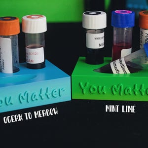 Puede incluir: Dos soportes rectangulares de plástico en azul y verde, cada uno con el texto "You Matter" en relieve. El soporte azul sostiene tres viales transparentes pequeños con tapas naranjas y blancas. El soporte verde sostiene tres viales con tapas de colores.