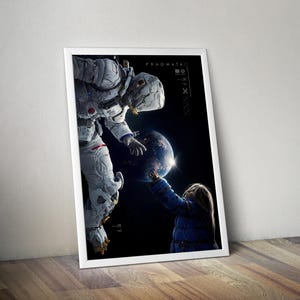 May include: Framed art print featuring an astronaut reaching towards a child touching a glowing Earth. The artwork has a dark background with the word "PRAGMATA" and other text. The astronaut is wearing a white spacesuit, and the child is wearing a blue jacket.