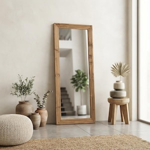 May include: A tall, rectangular floor mirror with a natural light brown wooden frame. The mirror reflects an interior space with a staircase and a potted plant. It is positioned upright against a plain wall, surrounded by various decorative items.