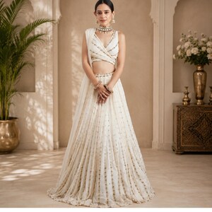 May include: A cream-colored lehenga set featuring a sleeveless wrap-style blouse and a floor-length skirt. Both pieces are adorned with vertical rows of metallic gold embroidery and small reflective sequins, creating a shimmering, textured appearance.
