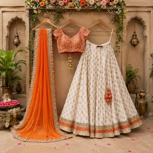 May include: A three-piece ensemble featuring an orange dupatta with a gold embroidered border, a short-sleeved orange blouse with intricate gold floral embroidery, and a cream-colored lehenga skirt with a gold pattern and an orange hem.