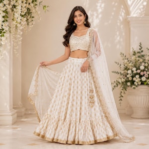 May include: A white and gold lehenga, a traditional Indian outfit, featuring a crop top and a long skirt. The skirt has a gold pattern and a ruffled hem. A sheer, white dupatta with gold trim completes the ensemble.