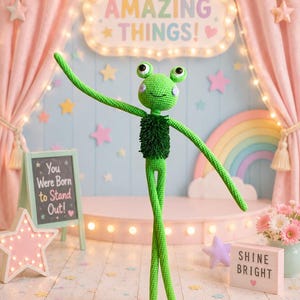 May include: A whimsical, green crocheted frog doll with long limbs and a cheerful expression stands on a pink stage. The background includes a sign that says "Let's Do AMAZING THINGS!" and "You Were Born to Stand Out!" with a rainbow and stars.
