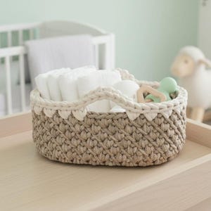 May include: An oval-shaped storage basket featuring a textured, tan-colored crocheted base and a thick, cream-colored crocheted rim. The basket is filled with several white folded cloths and a small, light green silicone ring-shaped toy.