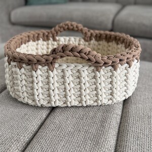 May include: An oval-shaped storage basket crafted from thick, chunky yarn. The main body of the basket features a vertical ribbed pattern in an off-white color, while the upper rim and two integrated handles are finished in a contrasting brown.
