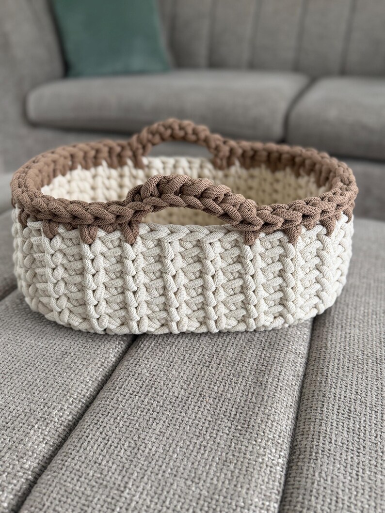 May include: An oval-shaped storage basket crafted from thick, chunky yarn. The main body of the basket features a vertical ribbed pattern in an off-white color, while the upper rim and two integrated handles are finished in a contrasting brown.