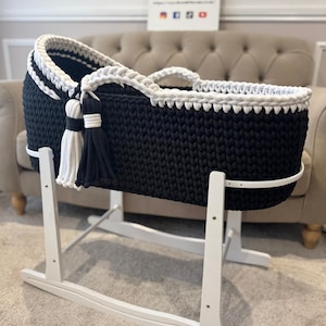 May include: A black and white knitted baby bassinet on a white wooden stand. The bassinet has a white braided trim and tassels. The stand is designed for rocking. The sign in the background says "The Little Lady".