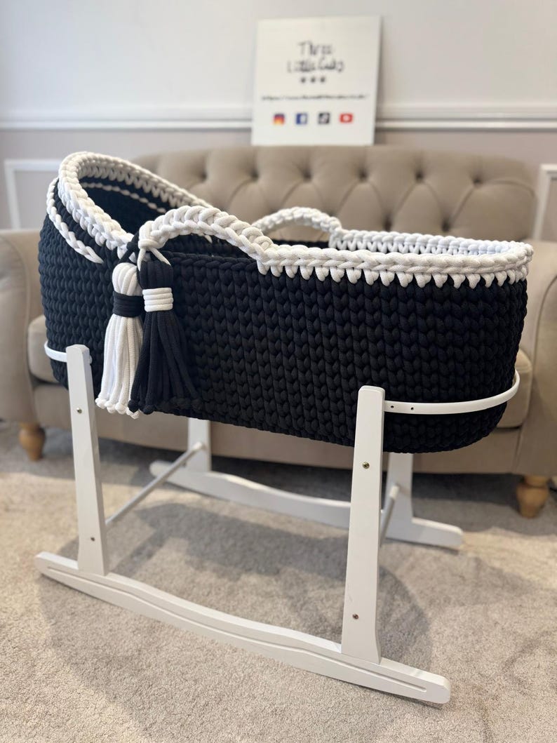May include: A black and white knitted baby bassinet on a white wooden stand. The bassinet has a white braided trim and tassels. The stand is designed for rocking. The sign in the background says "The Little Lady".