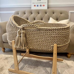 May include: A beige, hand-crocheted baby bassinet with a tassel detail, set on a natural wood rocking stand. The bassinet features a woven texture and a sturdy design. A sign in the background reads "Three Little Gals".