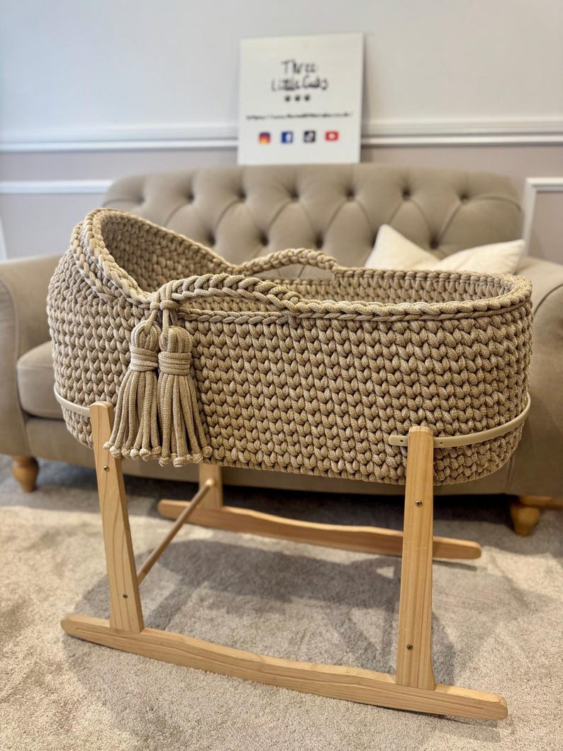 May include: A beige, hand-crocheted baby bassinet with a tassel detail, set on a natural wood rocking stand. The bassinet features a woven texture and a sturdy design. A sign in the background reads "Three Little Gals".