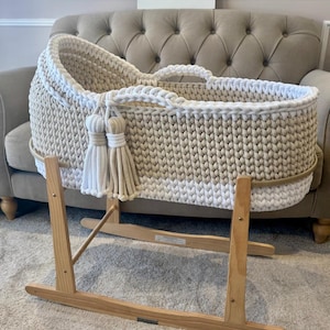 May include: A cream-colored, hand-knitted baby bassinet with a white trim and tassels. The bassinet sits on a wooden rocking stand. The sign in the background reads "Three Little Cubs".