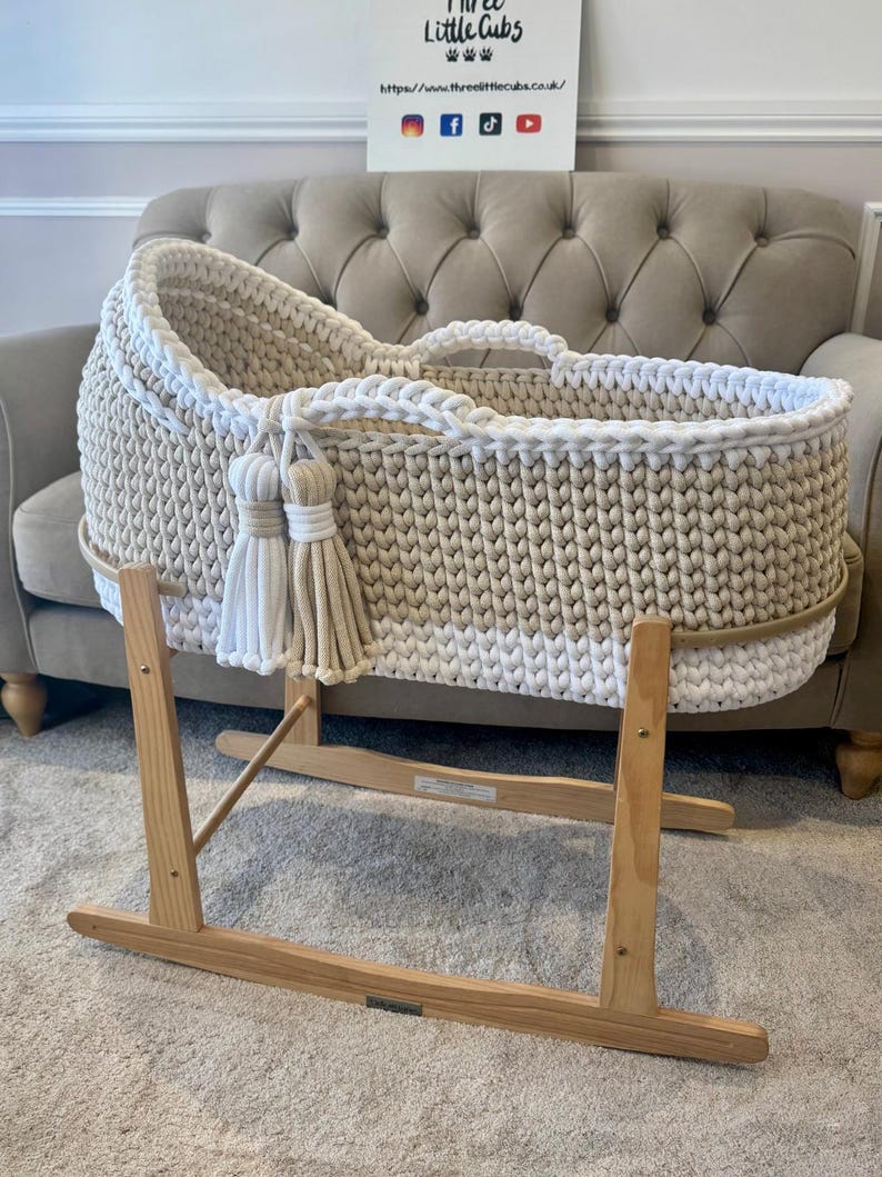 May include: A cream-colored, hand-knitted baby bassinet with a white trim and tassels. The bassinet sits on a wooden rocking stand. The sign in the background reads "Three Little Cubs".