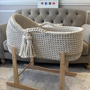 May include: A cream-colored, hand-knitted baby bassinet with a wooden rocking stand. The bassinet features a braided rim, tassels, and a handle. The wooden stand is light-colored. The sign in the background has the text "Three Little Cubs".