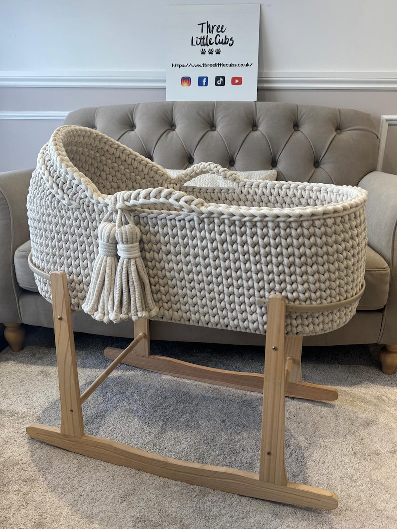 May include: A cream-colored, hand-knitted baby bassinet with a wooden rocking stand. The bassinet features a braided rim, tassels, and a handle. The wooden stand is light-colored. The sign in the background has the text "Three Little Cubs".