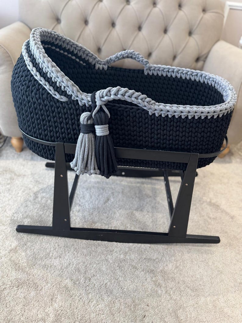 May include: A navy blue and gray crochet baby bassinet on a black rocking stand. The bassinet has a gray trim and two tassels in gray and black. The bassinet is designed for a baby.
